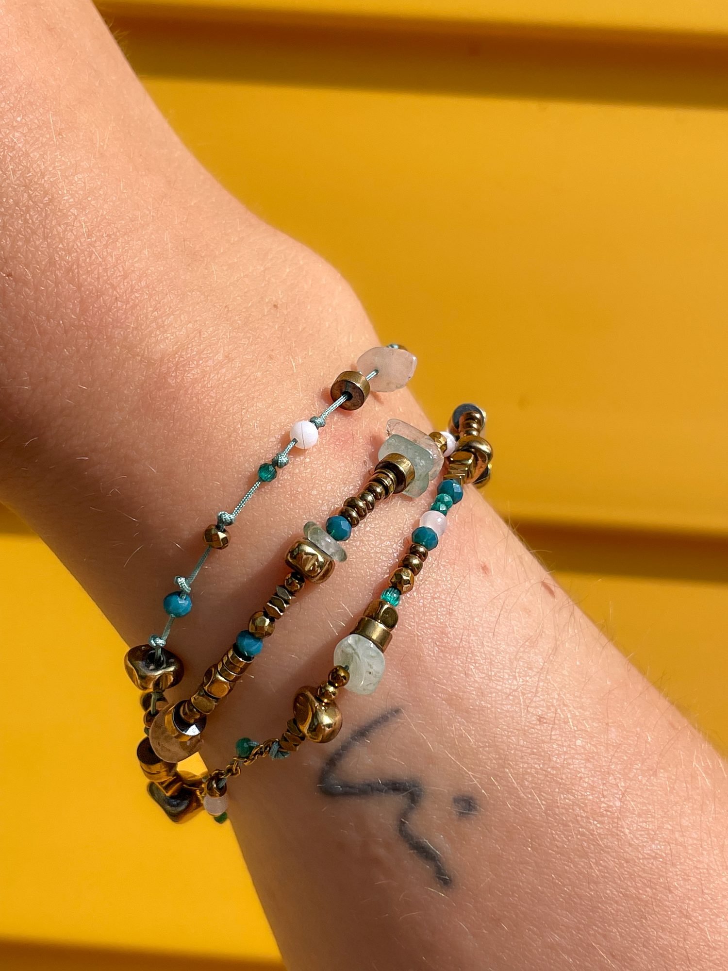 Trois bracelets ZouzBijoux en pierre naturelle et hématite dorée portés au poignet, devant un mur jaune
